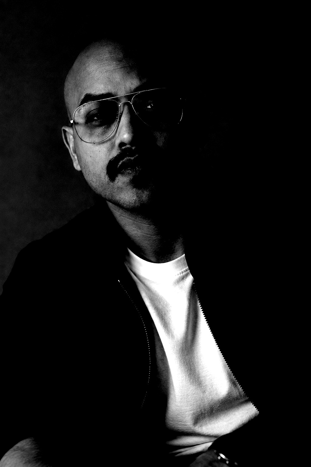 Andy Rozario, founder of Imagio Labs, in monochrome: a bald man in aviator glasses and a dark jacket over a white tee, lit from one side against a backdrop that has been crushed to black.